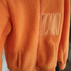 Orange fleecejacka från Peak Performance - Säljer en snygg orange fleecejacka från Peak Performance. Jackan har en dragkedja framtill och en praktisk bröstficka med dragkedja. Perfekt för kyliga dagar med sin mjuka och varma känsla. Broderad logga på ryggen ger en extra touch. Storlek small herrmodell. Använd enstaka gånger, som ny!
