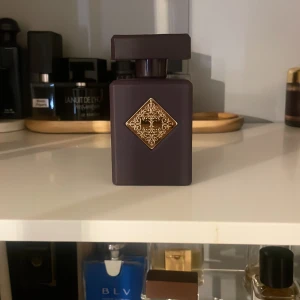 Initio side effect Parfym - Elegant parfymflaska från Initio med en mörklila färg och en dekorativ guldplatta på framsidan. Flaskan har en fyrkantig form med ett matchande lock. Perfekt för den som söker en lyxig doftupplevelse.