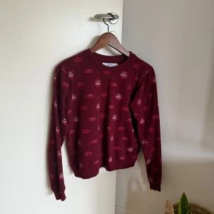 Vinröd sweatshirt med läppar från Dedicated - Vinröd sweatshirt från Dedicated i 100% ekologisk bomull. Tröjan har ett coolt mönster med röda läppar över hela och ribbade muddar vid ärmslut och hals. Perfekt för dig som vill sticka ut lite extra med din stil.