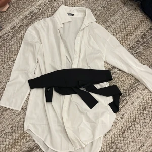 Vit skjorta med knytbälte  - Lång vit skjorta från SHEIN i storlek XS. Skjortan har klassisk krage, knappar framtill och långa ärmar. Medföljer ett brett svart knytbälte.