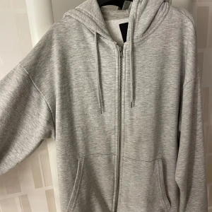 Ljusgrå hoodie med dragkedja 157 - Ljusgrå hoodie från 157 i storlek S. Tröjan har huva med snörning, hel dragkedja framtill och två stora fickor. Perfekt för chill dagar och enkel att matcha med jeans eller mjukisbyxor. Mjuk och skön bomullsblandning.