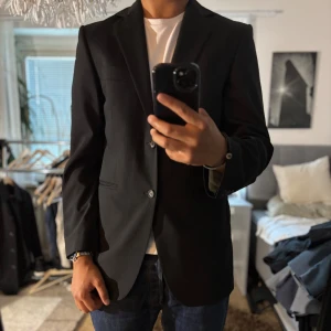 Svart randig kavaj Tiger of Sweden - Snygg svart kavaj från Tiger of Sweden i klassisk passform. Tunt randigt mönster, två knappar framtill och diskreta fickor. Tillverkad i en mix av ull och polyester med lite elastan för extra komfort. Perfekt för dig som vill ha en stilren och tidlös look.