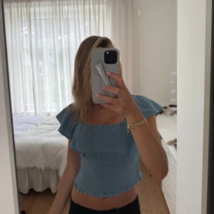 Blå smockad topp från Kappahl - Supersöt blå topp från Kappahl med smockad kropp och volangiga korta ärmar. Toppen har fyrkantig ringning och lätt croppad passform. Perfekt för varma dagar och ger en chill vibe till din outfit.