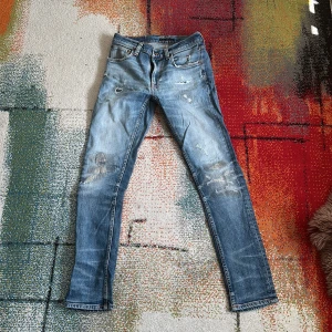 Nudie jeans - Säljer ett par ljusblå jeans från Nudie Jeans med slitna detaljer och lagningar på knäna. Klassisk femficksmodell med Nudie-logga på bakfickan och läderpatch i midjan. Jeansen har en rak passform och är tillverkade i bomull.