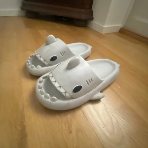 Grå hajformade sandaler - Unika sandaler i form av hajar, ljusgrå färg med svarta ögon och vita tänder. Tillverkade i mjuk syntet, perfekta för chill hemma eller på stranden. Rolig och lekfull design som sticker ut.