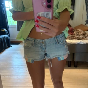 Ljusblåa jeansshorts från Hollister - Säljer ett par ljusblåa, lågmidjade jeansshorts från Hollister med slitna detaljer och fransig kant. Shortsen har klassiska regnbåge fickor, bälteshällor och en liten broderad logga. Insidan av benen har ett färgglatt rutigt tyg som syns under fransarna. Perfekta för sommaren! Mina favorit shorts:(
