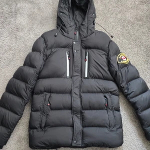 Svart dunjacka från Zavetti Canada - Säljer en svart dunjacka från Zavetti Canada med huva och feta vadderade partier. Jackan har flera dragkedjefickor framtill, snygga detaljer och en broderad patch på ärmen. Perfekt för kalla dagar och riktigt stilren look.