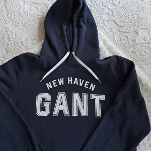 Marinblå hoodie från GANT New Haven - Snygg marinblå hoodie från GANT med vit text 'NEW HAVEN GANT' på bröstet. Klassisk huva med vita snören och mjukt, fleeceliknande foder på insidan. Perfekt för dig som gillar sportig och avslappnad stil.