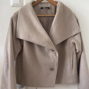 Beige kappa från Gina Tricot XS - Säljer en beige kappa från Gina Tricot i storlek XS. Jackan har en oversized krage, knäppning framtill med två knappar och långa ärmar. Materialet är mjukt och känns ulligt, perfekt för kyligare dagar. Enkel och stilren design som passar till många outfits.