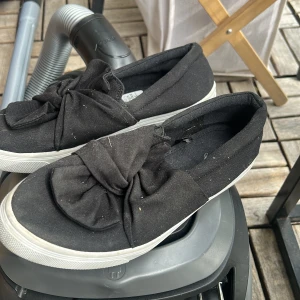 Svarta slip-on skor med rosett från Alley - Svarta slip-on skor från Alley med stor dekorativ rosett framtill. Skorna har vit platt sula och är tillverkade i textilmaterial. Perfekta för dig som gillar en enkel men ändå söt stil.