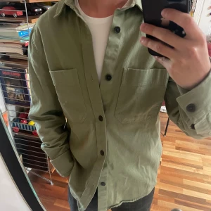 Grön overshirt - Olivgrön overshirt från H&M i bomull med två stora bröstfickor och svarta knappar. Skjortan har klassisk krage och lång ärm, perfekt att bära öppen över en t-shirt för en avslappnad look. 