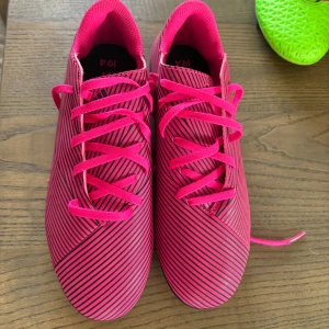 Adidas rosa fotbollsskor med svarta ränder - Snygga fotbollsskor från Adidas i starkt rosa med svarta ränder och tunna svarta linjer över hela skon. Skorna har snörning och är designade för att ge bra grepp på planen. Perfekta för dig som vill sticka ut på matchen.