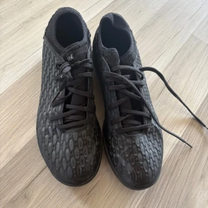 Svarta fotbollsskor med mönstrad yta - Säljer ett par svarta fotbollsskor med coolt, strukturerat mönster på ovandelen. Skorna har snörning och låg profil, perfekt för snabba rörelser på planen. Ovandelen är i syntetmaterial och sulan är platt för bra grepp inomhus.