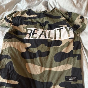 Camouflage t-shirt från Woxo Reality - Cool t-shirt från Woxo med grön, beige och svart camouflage-mönster och stor texten 'REALITY' framtill. Skön passform och tillverkad i mjuk bomull. Perfekt för dig som gillar streetstyle och vill sticka ut lite extra.