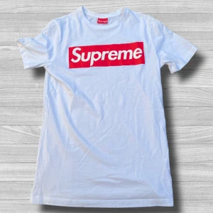 Vit Supreme t-shirt  - Vit t-shirt från Supreme med den ikoniska röda box logon framtill. Klassisk rund halsringning och korta ärmar. Tillverkad i mjuk bomull, perfekt för en clean streetwear-look.