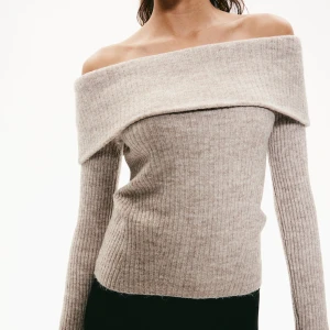 Beige offshoulder stickad tröja - Superfin beige stickad tröja med ribbad struktur och bred offshoulder-krage. Långärmad och mjuk, perfekt för lager på lager. Snygg till både jeans och kjol, ger en chill men ändå trendig vibe.