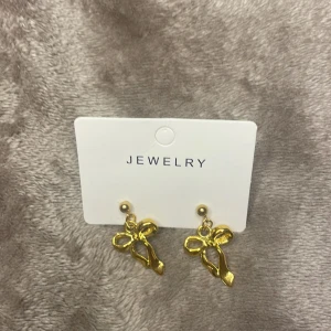 Guldiga örhängen med rosett - Supersöta örhängen i guld med hängande rosettform. De har en blank finish och är perfekta för att ge din look en extra touch av stil. Säljs på originalkortet, enkla att matcha med andra smycken.