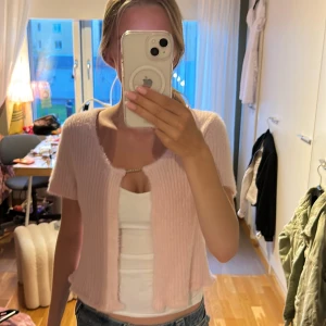 Ljusrosa stickad kofta från Kendy - Supergullig ljusrosa stickad kofta från Kendy med kort ärm och croppad passform. Koftan har ribbad struktur, rund halsringning och stängs framtill med en liten dekorativ brosch. Perfekt att slänga över ett linne för en söt look.