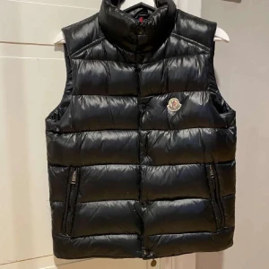 Moncler väst - Snygg svart dunväst från Moncler med hög krage och två dragkedjefickor framtill. Västen har quiltad design och klassisk Moncler-logga på bröstet. Perfekt att slänga över en hoodie eller tröja för en clean look.
