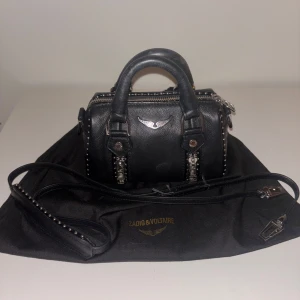 Zadig & Voltaire väska - handväska från Zadig & Voltaire i äkta skinn med silverfärgade detaljer, nitar och dragkedjor. Väskan har både handtag och avtagbar axelrem, samt ikonisk logga framtill. Perfekt storlek för det viktigaste och ger en edgy vibe till din outfit. De e den minsta storleken💕