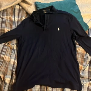 Marinblå pikétröja från Polo Ralph Lauren - Klassisk marinblå långärmad pikétröja från Polo Ralph Lauren med ikonisk broderad logga på bröstet. Tröjan har krage och knäppning framtill, tillverkad i mjuk bomull som känns skön mot huden. Perfekt för en stilren och avslappnad look.