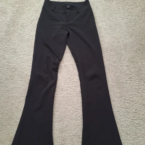 Svarta bootcut byxor Gina Tricot S - Svarta bootcut byxor från Gina Tricot i storlek S. Byxorna har hög midja, två knappar framtill, bälteshällor och bakfickor med lock och knapp. Snygg passform med utsvängda ben, perfekta till sneakers eller boots.