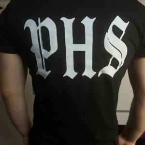 Svart PHS Support t-shirt i bomull - Svart t-shirt fràn PHS med stor vit logga pà ryggen och mindre tryck med knogjärn och texten 'SUPPORT' pa bröstet. Klassisk rund hals och korta ärmar. Tillverkad i mjuk bomull, perfekt för dig som gillar streetwear vibes.