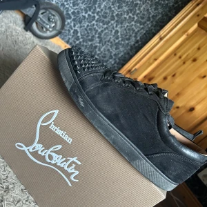Christian Louboutin svarta sneakers med spikar  - Svarta sneakers från Christian Louboutin i mocka med ikoniska röda sulor. Skorna har snörning och coola svarta nitar på tån för en edgy vibe. Låg modell med rund tå och diskreta metalldetaljer på sidan. Perfekta för dig som vill sticka ut.