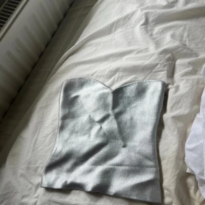 Silver axelbandslös topp från Zara - Säljer en metallic silver axelbandslös topp från Zara. Toppen har en snygg hjärtformad urringning och sitter tight över överkroppen. Materialet är glansigt och ger en futuristisk vibe. Perfekt att styla med jeans eller kjol för en cool look.