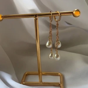 Guldiga örhängen med pärldroppar - Snygga guldiga örhängen med tunna kedjor och hängande droppformade pärlor. Perfekta för att ge en elegant touch till din look. Örhängena har en stilren design med glansig finish och vita pärlor som fångar ljuset fint.