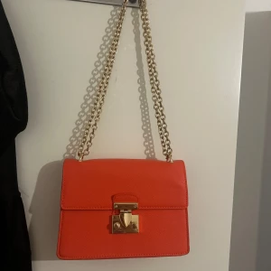 Orange axelväska med guldkedja - Snygg orange axelväska med stilren, fyrkantig form och guldfärgad kedjerem. Väskan har ett elegant lås framtill i guld och är tillverkad i ett slätt, strukturerat material. Perfekt för att lyfta vilken outfit som helst.