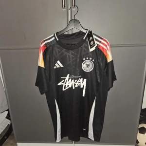 Tysklands Stüssy x Adidas fotbollströja XL - Snygg svart Tyskland fotbollströja i samarbete mellan Adidas och Stüssy. Tröjan har korta ärmar, vita och röda detaljer på axlarna, samt Tysklands landslagsemblem och Stüssy-logga på bröstet. Tillverkad i lätt och ventilerande material, perfekt för match eller träning.