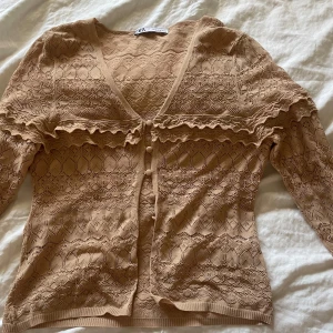 Beige spetskofta med volang från Zara - Storlek m men passar allt från xs -m. Supersöt beige kofta från Zara i spets med v-ringning, knappar framtill och volangdetaljer längs bröstet och ärmsluten. Långärmad och figurnära passform.