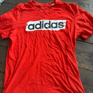 Röd Adidas t-shirt med logga - Röd t-shirt från Adidas med stor svartvit logga på bröstet. Klassisk rund halsringning och korta ärmar. Skön och lätt i bomull, perfekt för en avslappnad stil.