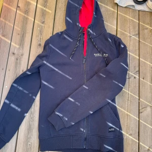 Mörkblå hoodie Race Marine S - Mörkblå hoodie från Race Marine med röd insida på huvan och vita tryckdetaljer. Dragkedja framtill, snörning i huvan och broderad siffra 83 på ärmen. Tillverkad i mjuk bomull och polyesterblandning.