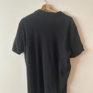 Svart pikétröja från Ralph Lauren - Klassisk svart pikétröja från Ralph Lauren med vit broderad logga på bröstet. Tröjan har korta ärmar, krage och två knappar framtill. Tillverkad i mjuk bomull som känns skön mot huden. Perfekt för en clean och stilren look.