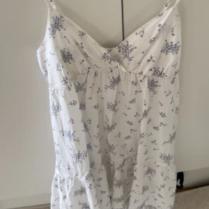 Vit blommig sommarklänning från Nelly - Supersöt vit klänning från Nelly med tunna axelband och blått blommönster. Lätt och luftig modell med v-ringning och volang nedtill, perfekt för varma dagar. Sitter skönt och har en romantisk vibe.
