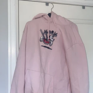 Rosa hoodie med Mickey Mouse-brodyr - Säljer en ljusrosa hoodie med broderad Mickey Mouse på bröstet. Tröjan har huva med snörning och en stor magficka. Perfekt för dig som gillar Disney och vill ha en chill och snygg stil.