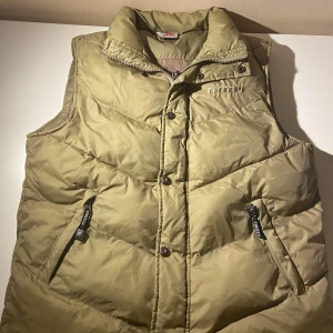 Beige dunväst från Firefly - Snygg beige dunväst från Firefly med hög krage och dragkedja samt knappar framtill. Västen har två fickor med dragkedja och broderad logga på bröstet. Perfekt för lager-på-lager och chillare dagar.