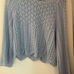 Ljusblå stickad tröja med spetsmönster - Superfin ljusblå stickad tröja med spetsmönster och vågig nederkant. Tröjan har långärmade ärmar och ett luftigt, detaljerat hålmönster som ger en romantisk och trendig vibe. Perfekt att styla med jeans eller kjol för en chill look.