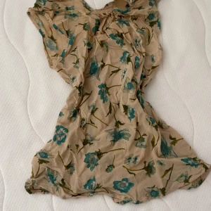 Blommig blus från Nygårds Anna - Supersöt blus från Nygårds Anna Bengtsson i beige med blågröna blommor. Klänningen har en lätt och luftig känsla, korta ärmar och v-ringning. Perfekt för varma dagar.