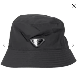 Svart bucket hat från Prada - Svart bucket hat från Prada med ikonisk metallogo framtill. Klassisk form med bredare brätte och sydda detaljer runt om. Tillverkad i glansig syntet som ger en clean och modern vibe. Perfekt accessoar för att lyfta din streetstyle.