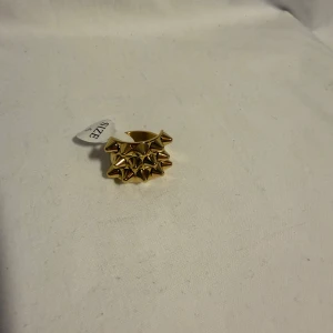 Guldig ring med nitar, storlek 6 - Cool statementring i guldton med bred design och flera spetsiga nitar framtill. Perfekt för dig som vill sticka ut och ge din look en edgy vibe. Ringen är i storlek 6 och har en blank finish som verkligen fångar ögat.