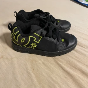 Svarta DC sneakers med neonlogga - Svarta sneakers från DC Shoes med stor neonfärgad DC-logga på sidan och detaljer i gult. Skorna har snörning, platt sula och är tillverkade i skinn och syntet. Perfekta för dig som gillar streetstyle och skate.
