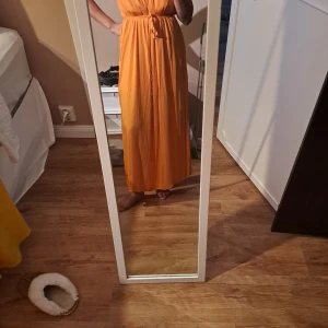 Orange långklänning med knyt i midjan - Säljer en snygg orange långklänning med smala axelband och knyt i midjan. Klänningen har en luftig och flowy passform, perfekt för varma dagar. Enkel och stilren design som ger en fräsch look. Sidenliknande material.