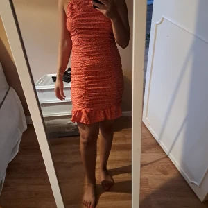 Röd kort volangklänning med smala axelband - Supersöt röd kort klänning med smala axelband och volang nertill. Klänningen har rynkade detaljer över hela och ett litet vitt mönster som ger en fräsch sommarkänsla. Sitter tight och framhäver figuren på ett snyggt sätt.