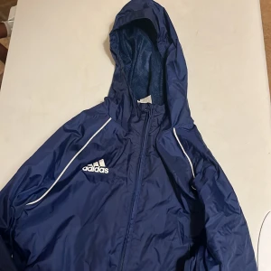 Adidas Vindjacka - Snygg marinblå vindjacka från Adidas med vit logga på bröstet och vita detaljer längs axlarna. Jackan har huva, dragkedja framtill och elastiska muddar vid ärmsluten. Perfekt för blåsiga dagar och sportiga outfits.