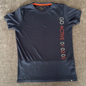 Mörkblå sportig t-shirt Go Active - Mörkblå tränings t-shirt från Go Active med orange detaljer och tryck längs sidan. Tillverkad i stretchigt funktionsmaterial med meshpanel nedtill för extra ventilation. Perfekt för träning eller sportiga dagar.