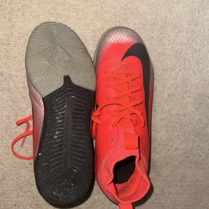 Nike Mercurial röda fotbollsskor - Snygga Nike Mercurial fotbollsskor i rött med svarta detaljer och silvermönster på sidorna. Skorna har snörning, högre skaft och en platt sula som passar perfekt för spel på konstgräs eller inomhus. Materialet är syntet och skon har en sportig vibe.