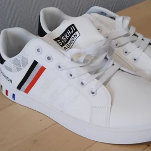 Vita sneakers med färgdetaljer från Fashion Sport - Fräscha vita sneakers från Fashion Sport med coola röda, svarta och blå detaljer på sidan och sulan. Skorna har snörning och en sportig vibe, perfekt för dig som gillar stilrena men ändå unika skor. Tillverkade i syntetmaterial.